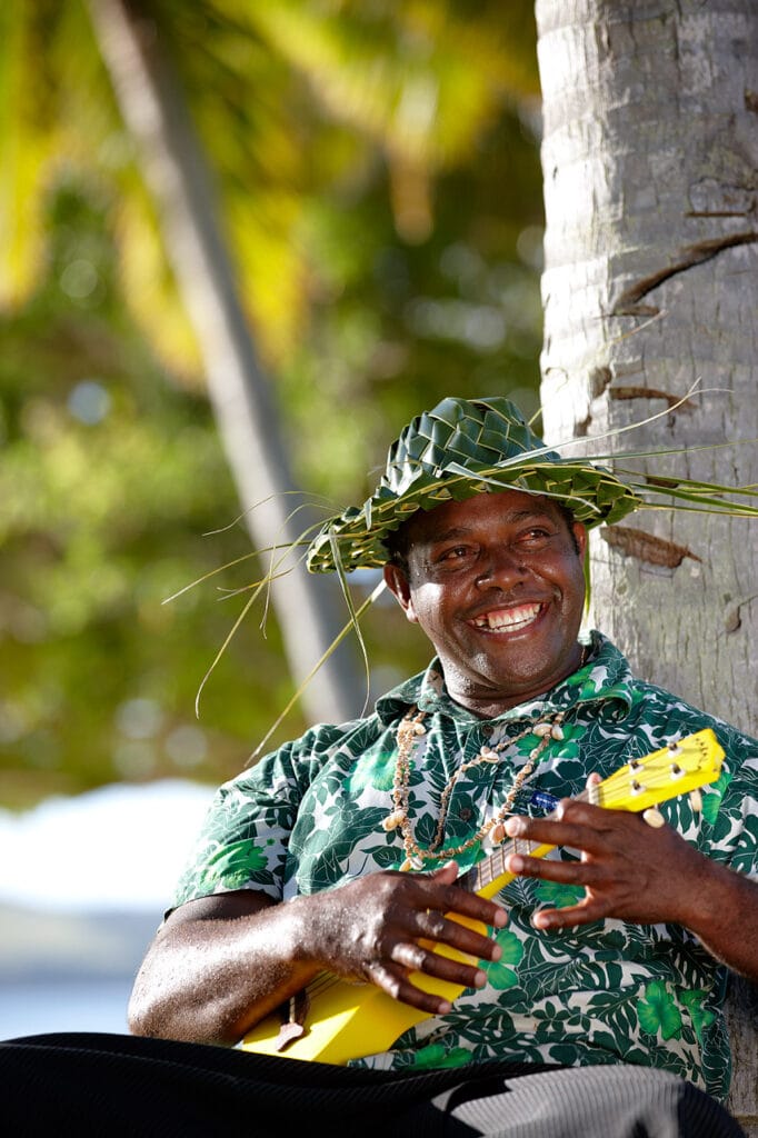 Smiling man in leaf hat playing a yellow ukulele by a palm tree.