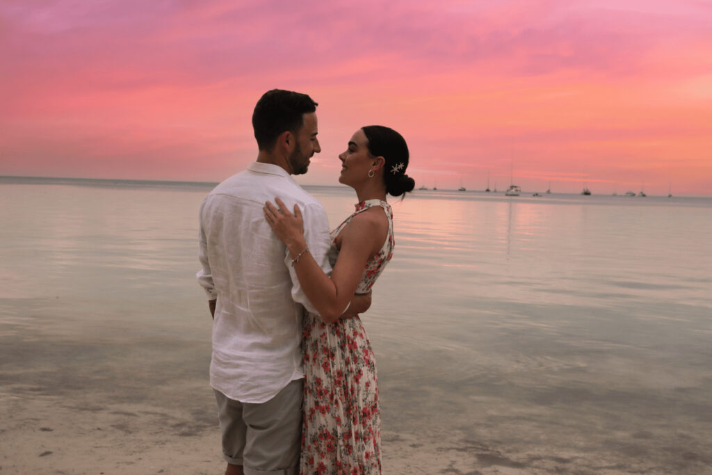 Couple embracing at sunset on the beach with sailboats in the distance.