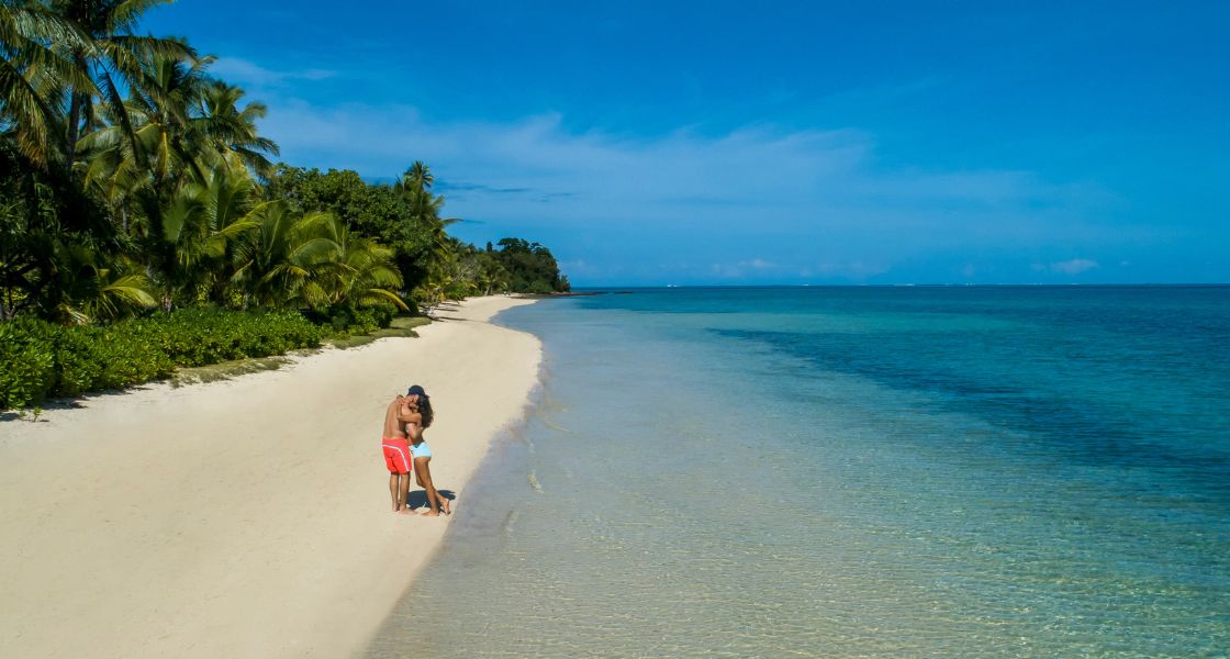 Couple in swimsuits hugging on a long powdery beach with clear turquoise lagoon.